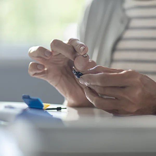 Close up of an older woman's hands holding her hearing aid in one hand and a small hearing aid battery in the other as she carefully replaces her hearing aid battery