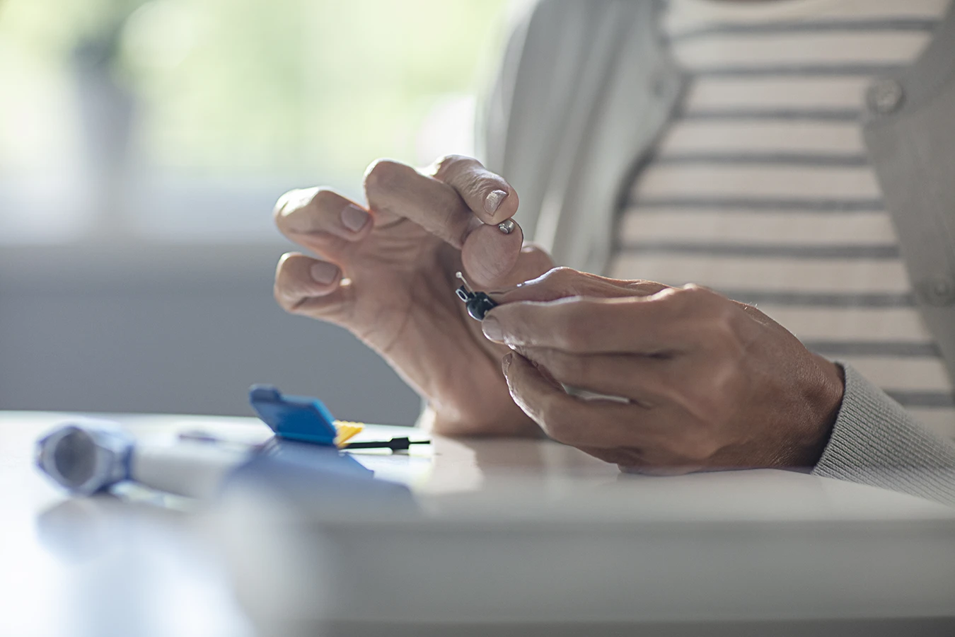Close up of an older woman's hands holding her hearing aid in one hand and a small hearing aid battery in the other as she carefully replaces her hearing aid battery