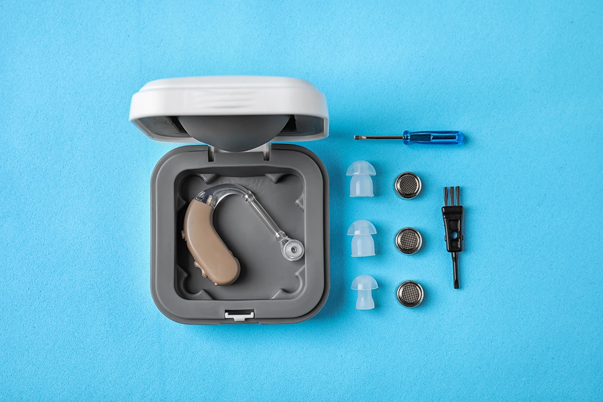 A BTE hearing aid in a case with hearing aid cleaning supplies placed next to it on a blue background