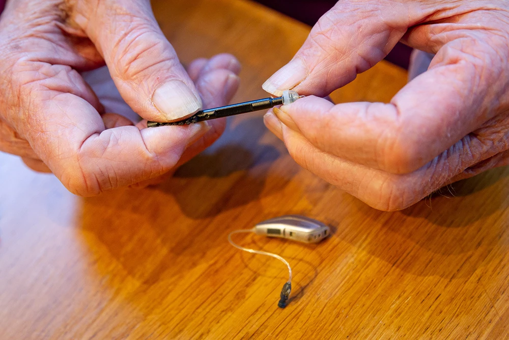 Mature male hands cleaning a BTE hearing aid above a wooden surface.