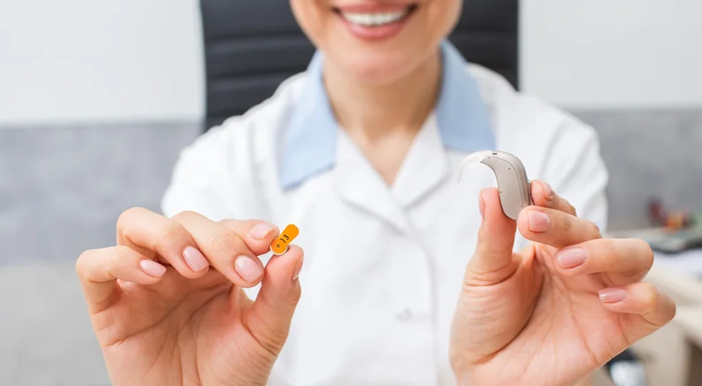 A hearing care specialist holding up a BTE hearing aid and an Orange , size 13 hearing aid battery