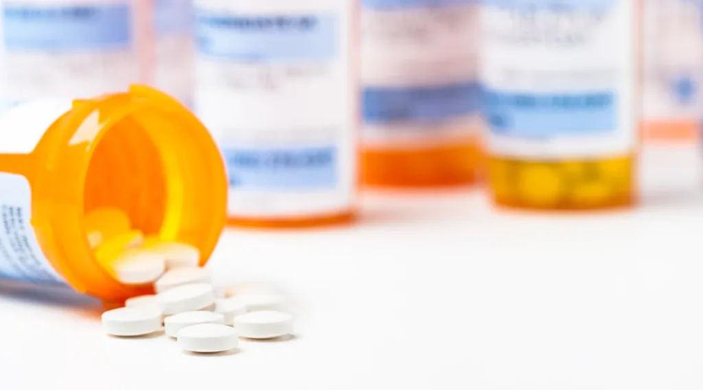 Round white prescription medicine pills spilling from a medication bottle with numerous full bottles in background