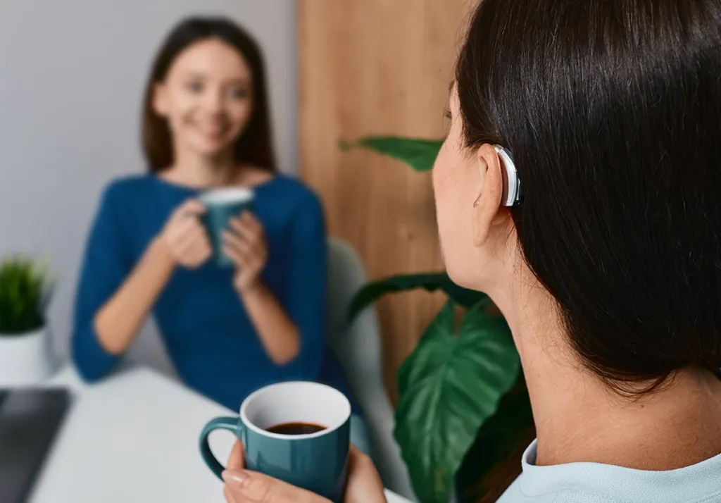A young women wearing a hearing aid, sitting in a cafe with her friend chatting, demonstrating how hearing aids enhance speech clarity even in challenging environments