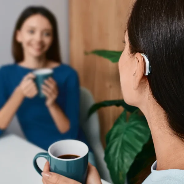 A young women wearing a hearing aid, sitting in a cafe with her friend chatting, demonstrating how hearing aids enhance speech clarity even in challenging environments