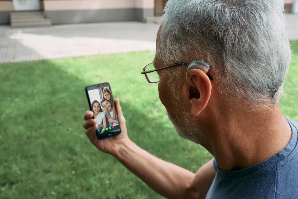 An older man using his Bluetooth hearing aids with his smartphone to facetime with his family.