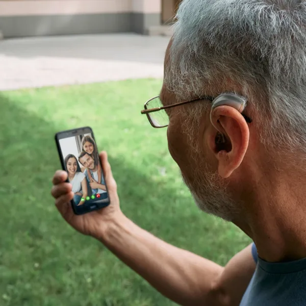 An older man using his Bluetooth hearing aids with his smartphone to facetime with his family.