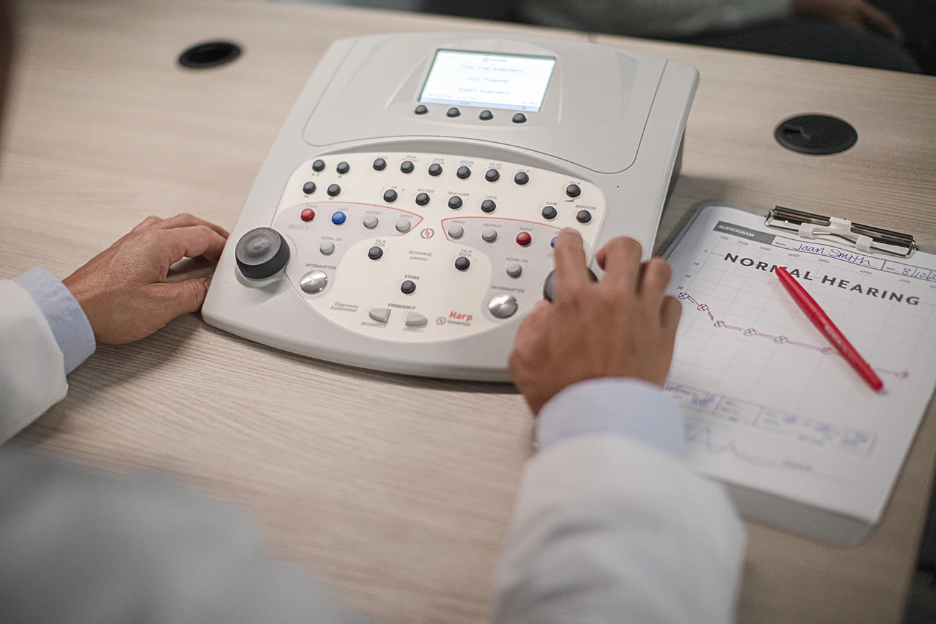 A hearing care specialist using a The device is an Inventis Harp diagnostic audiometer used for performing air, bone, and speech hearing tests, with a clipboard next to hime with text that reads, "NORMAL HEARING"