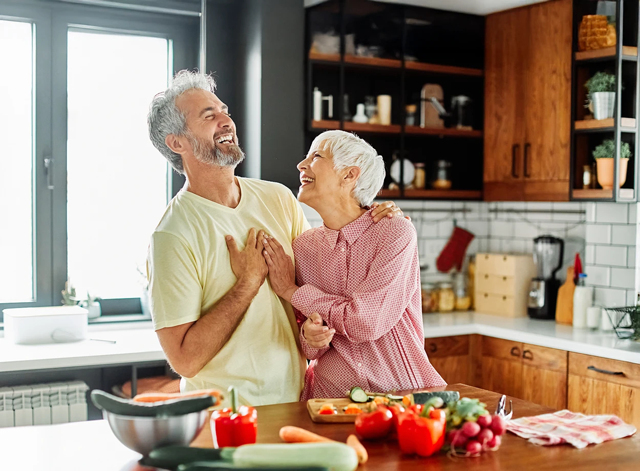 Older woman and man, married couple, happily preparing food together in the kitchen, cooking a healthy meal together.