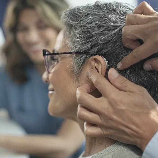 The side profile of a woman in an a hearing clinic during a hearing aid consultation, as her hearing care specialists fits a new hearing aids in her ear
