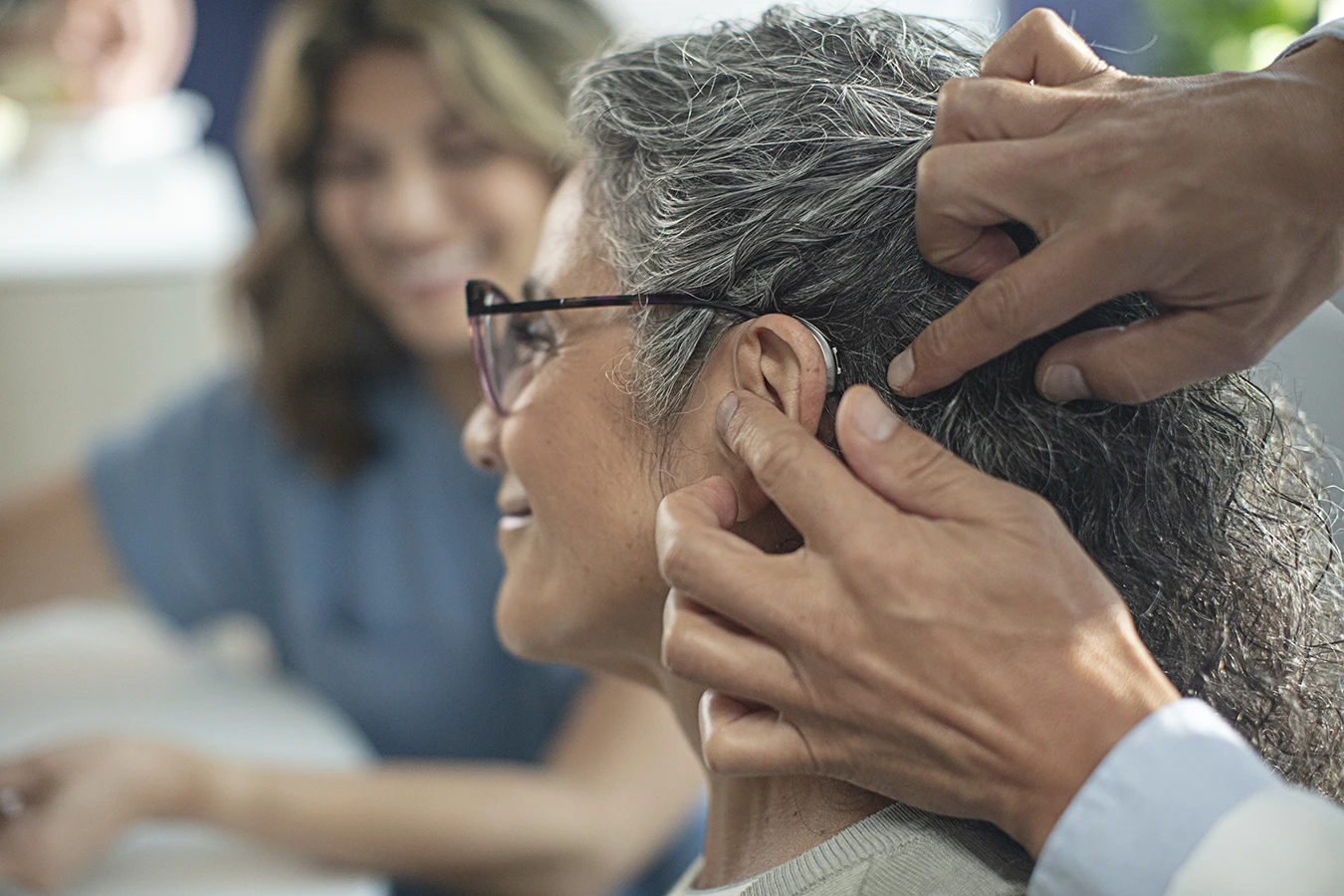 The side profile of a woman in an a hearing clinic during a hearing aid consultation, as her hearing care specialists fits a new hearing aids in her ear