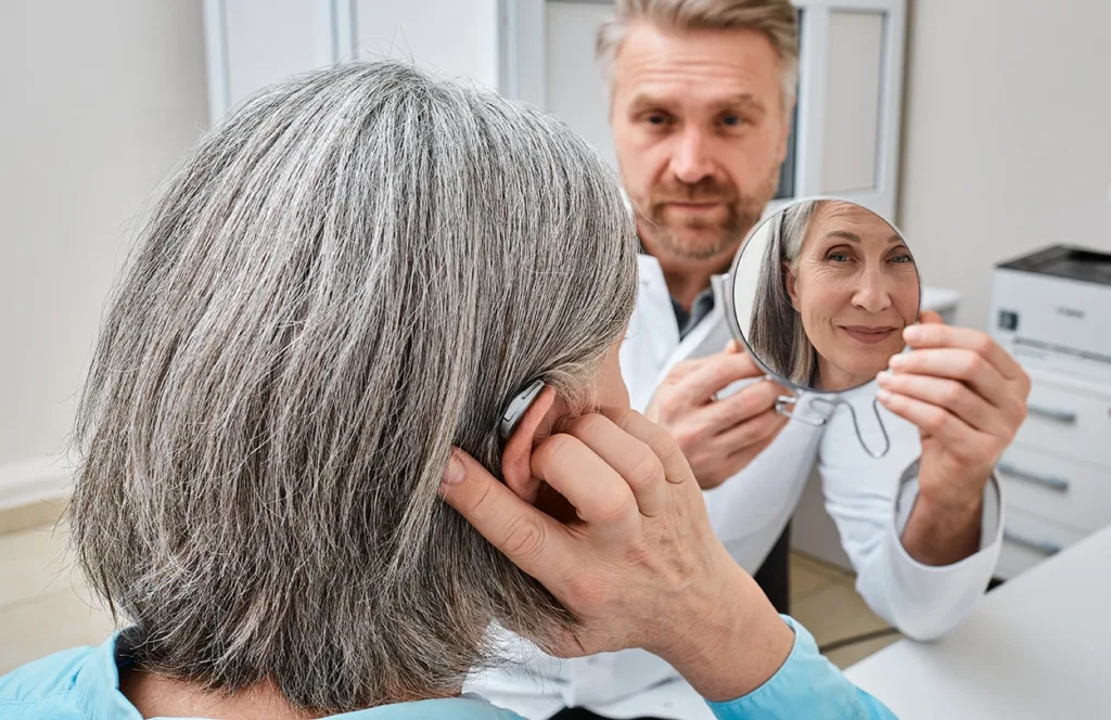 Older adult woman with severe hearing loss trying on a hearing aid while looking into a mirror held by a hearing aid specialist in a hearing loss care clinic.