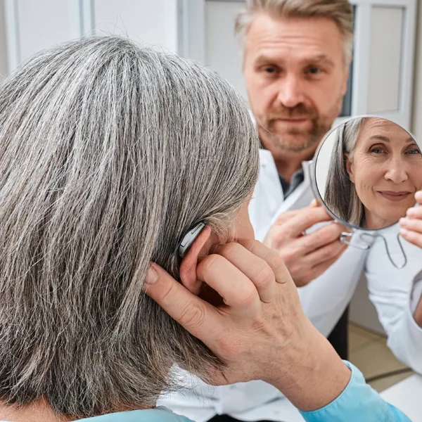 Older adult woman with severe hearing loss trying on a hearing aid while looking into a mirror held by a hearing aid specialist in a hearing loss care clinic.