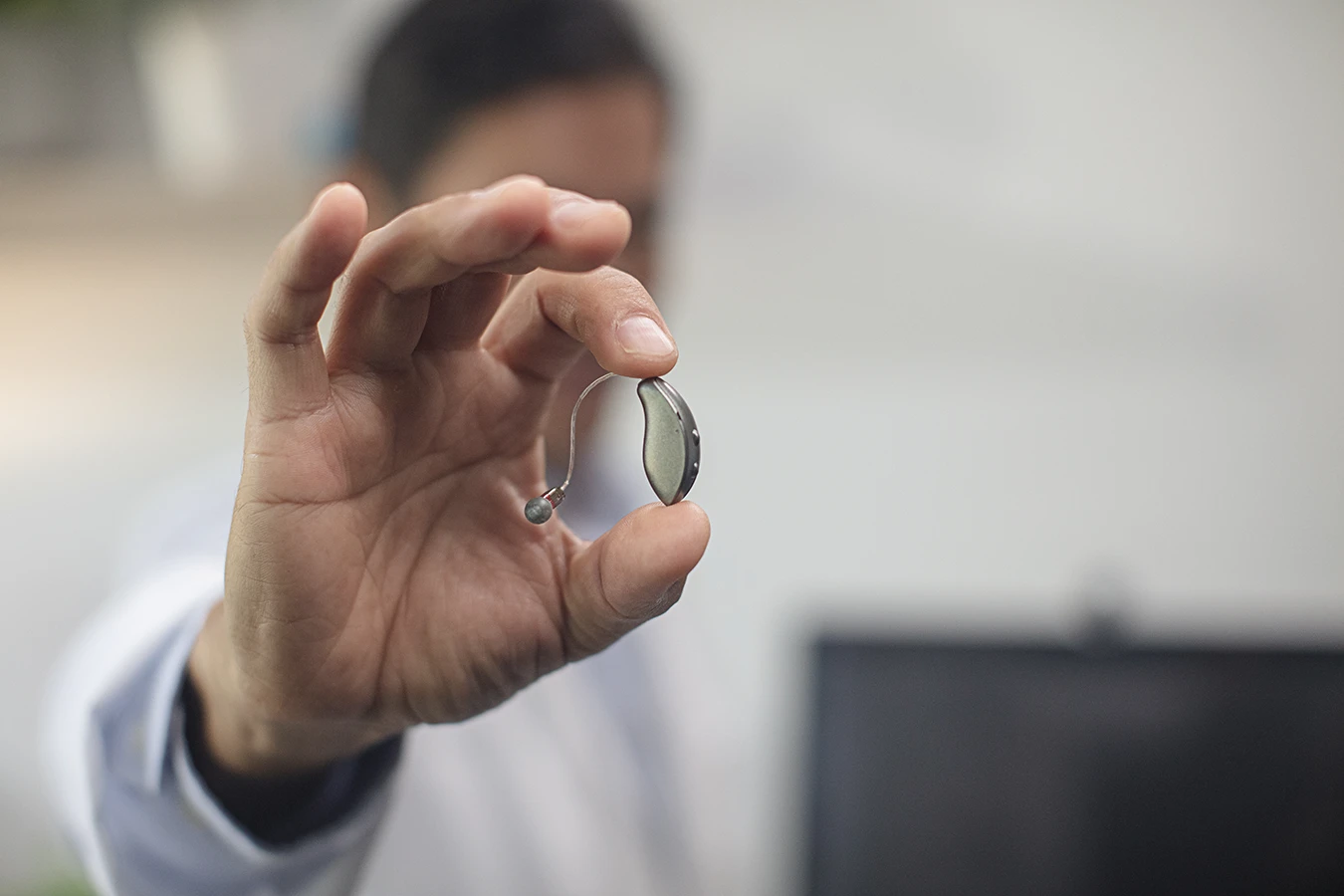 A close up of a behind-the-ear BTE hearing aid being held up by a hearing care specialist who is blurry in the background