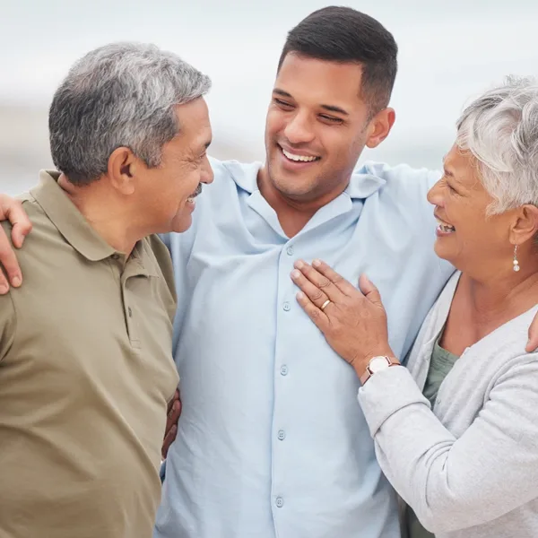 An adult man with his senior parents standing together, hugging, smiling, and laughing.