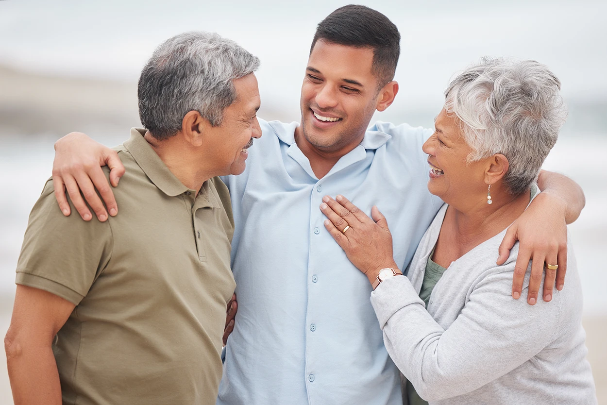 An adult man with his senior parents standing together, hugging, smiling, and laughing.