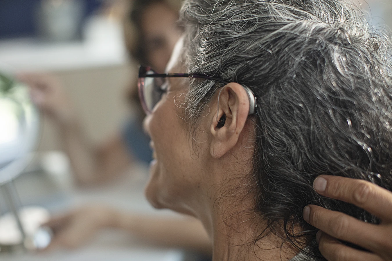 The side profile of a woman in an a hearing clinic during a hearing aid consultation, as her hearing care specialists fits a new hearing aids in her ear