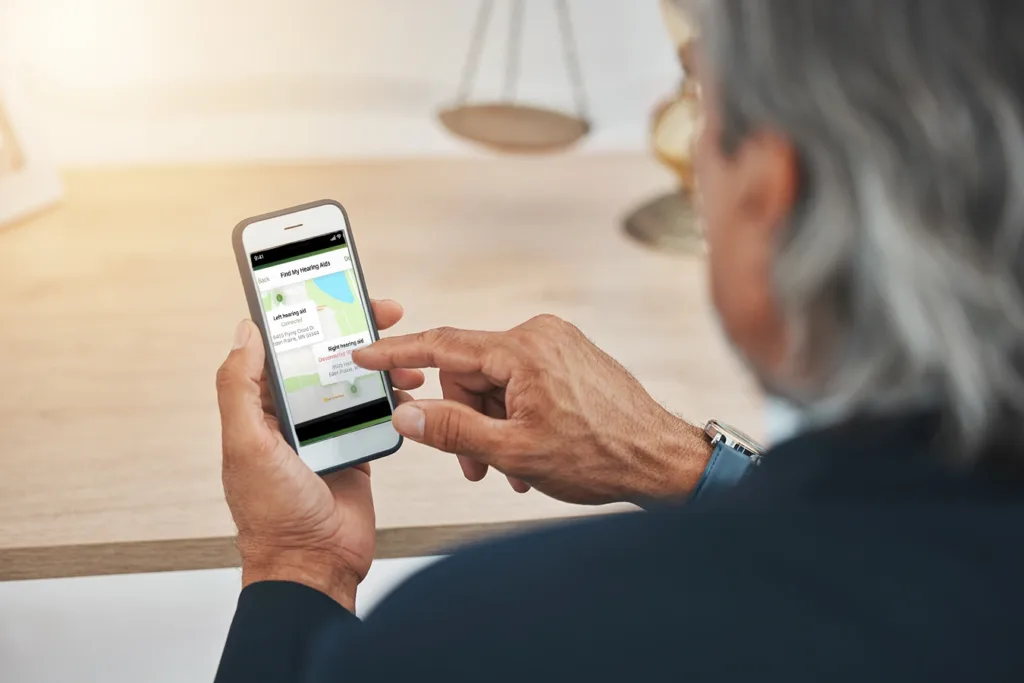 An older man with grey hair looking down at his smartphone, showing the Find My Hearing Aid app tool available in both the My Audibel and Thrive Hearing Control hearing aid apps and compatible with all Audible brand Bluetooth hearing aids, allowing him to track a lost hearing aid with ease.
