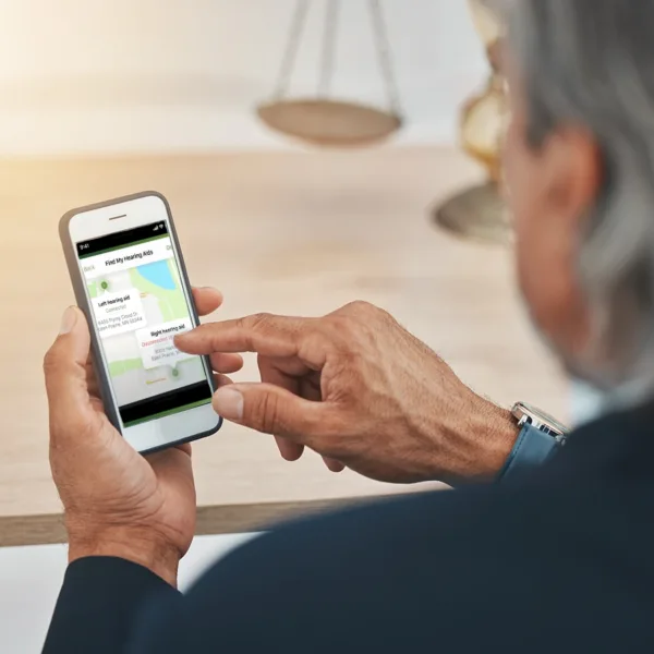 An older man with grey hair looking down at his smartphone, showing the Find My Hearing Aid app tool available in both the My Audibel and Thrive Hearing Control hearing aid apps and compatible with all Audible brand Bluetooth hearing aids, allowing him to track a lost hearing aid with ease.