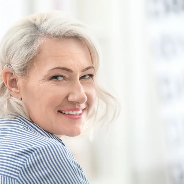 An older adult women with grey hair, wearing a behind the ear hearing aid, smiling and looking at the camera