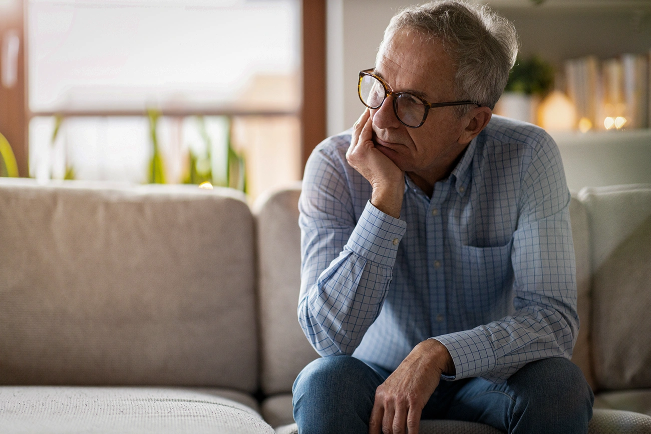 A senior man sitting alone in his home looking worried, sad, and tired due to listening fatigue from untreated hearing loss.
