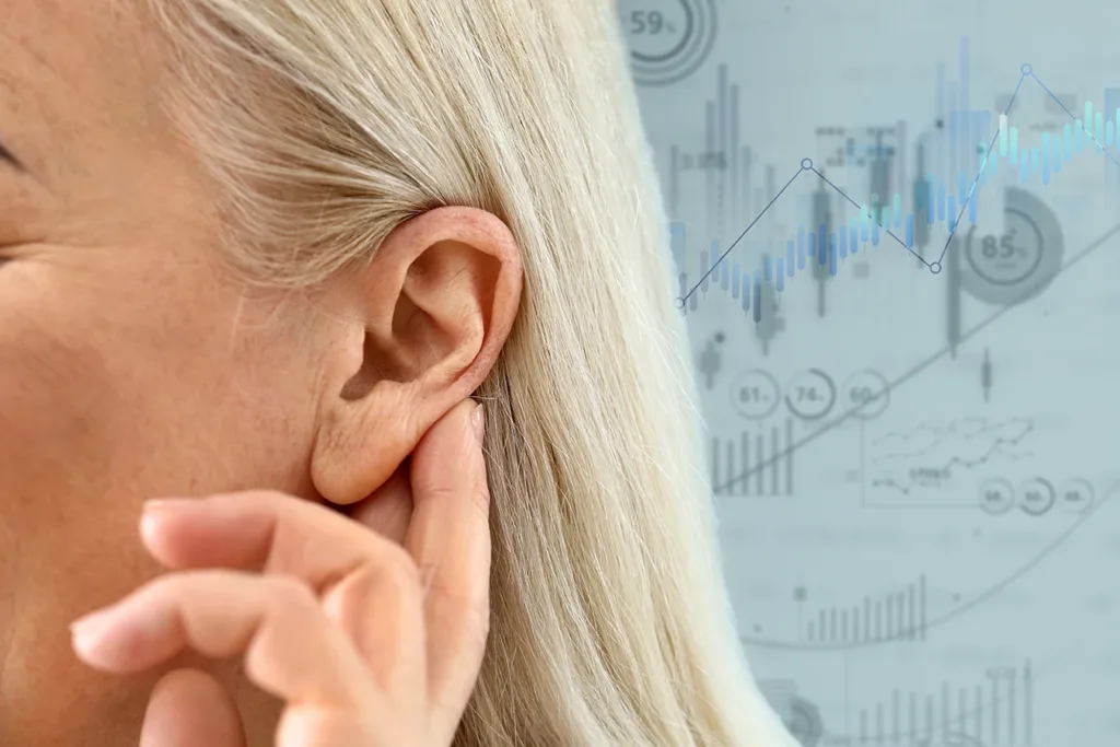 The side profile of a woman holding her hand up to her ear as if to help her hear better, with statistic graphics out of focus in the background, showing the concept of hearing loss statistics in the United States