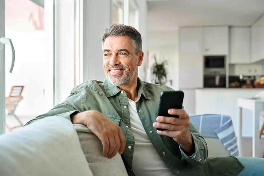 Happy mature middle aged man holding smartphone sitting at home on sofa. Symbolizes the latest hearing aid technology and Bluetooth Connectivity and Streaming Technology