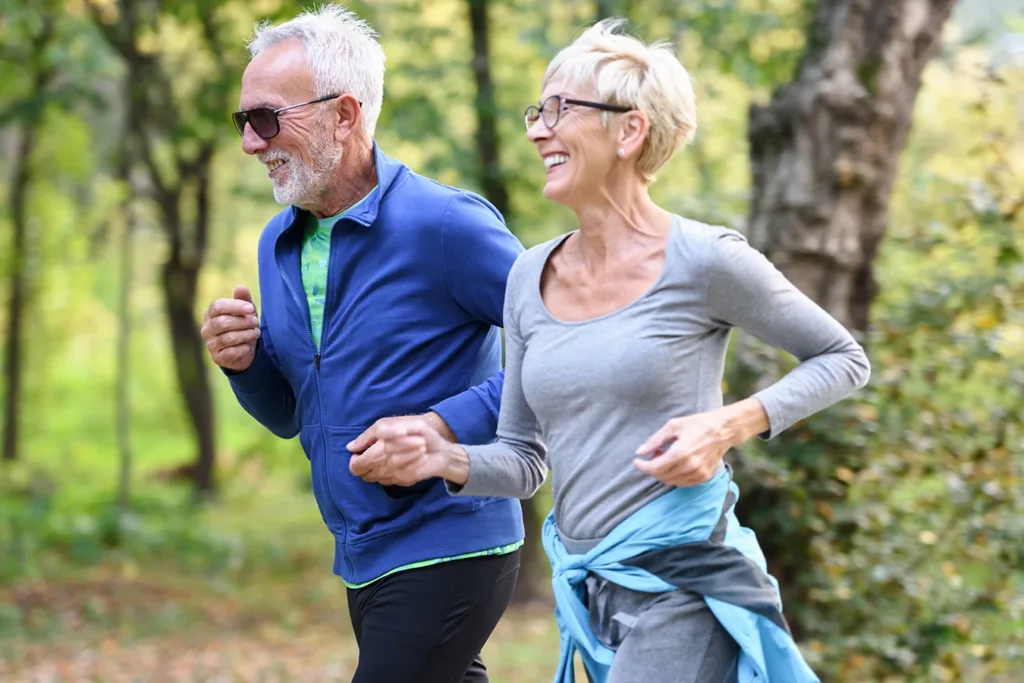 A happy retired couple, husband and wife, older adults, over the age of 60, dressed in workout clothes, jogging outside together through a beautiful, wooded park