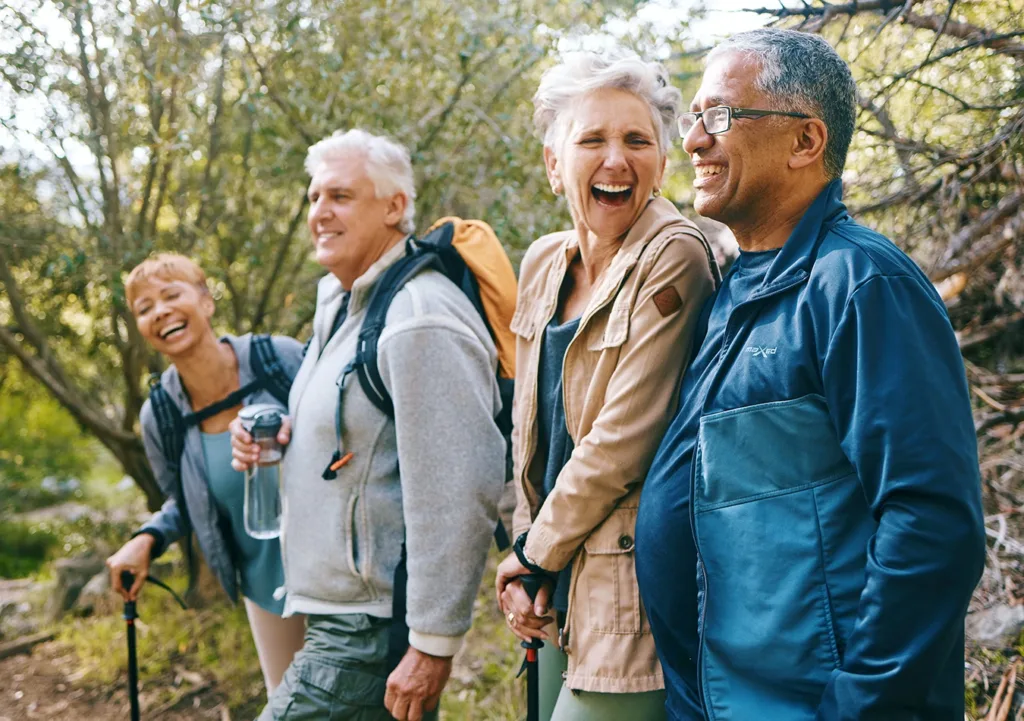 Four older adults, men and women, dressed in outdoorsy clothings, laughing, smiling, and talking as they hike together through a wooded area.