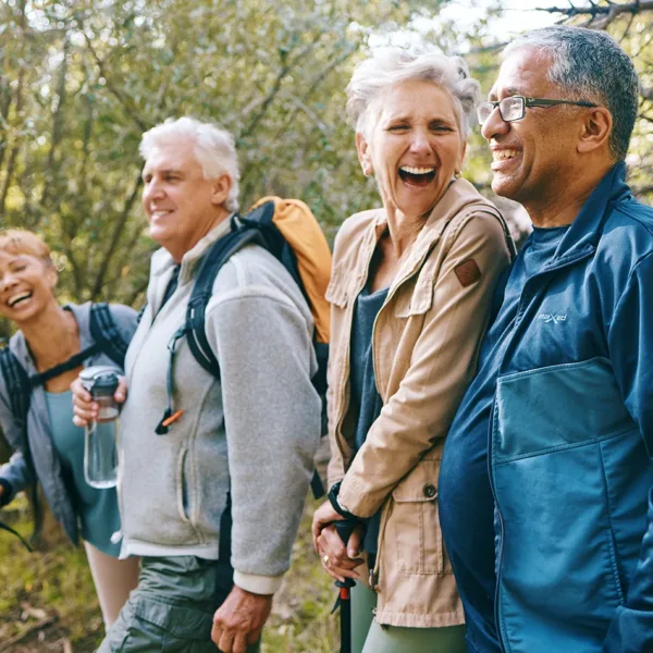 Four older adults, men and women, dressed in outdoorsy clothings, laughing, smiling, and talking as they hike together through a wooded area.