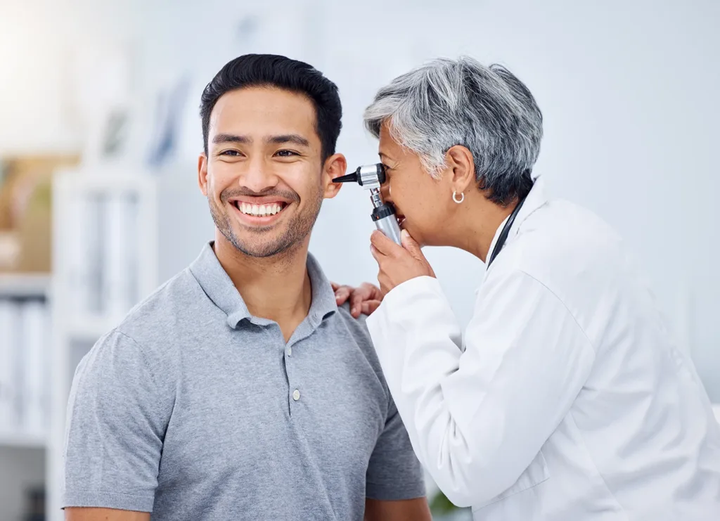 A young adult man at a hearing clinic, getting a hearing test by a hearing care specialist, looking into his ear with an otoscope