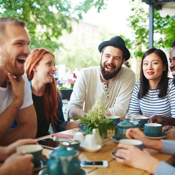 A group of seven young adults, men and women, sitting at a cafe table together talking, smiling, and laughing.