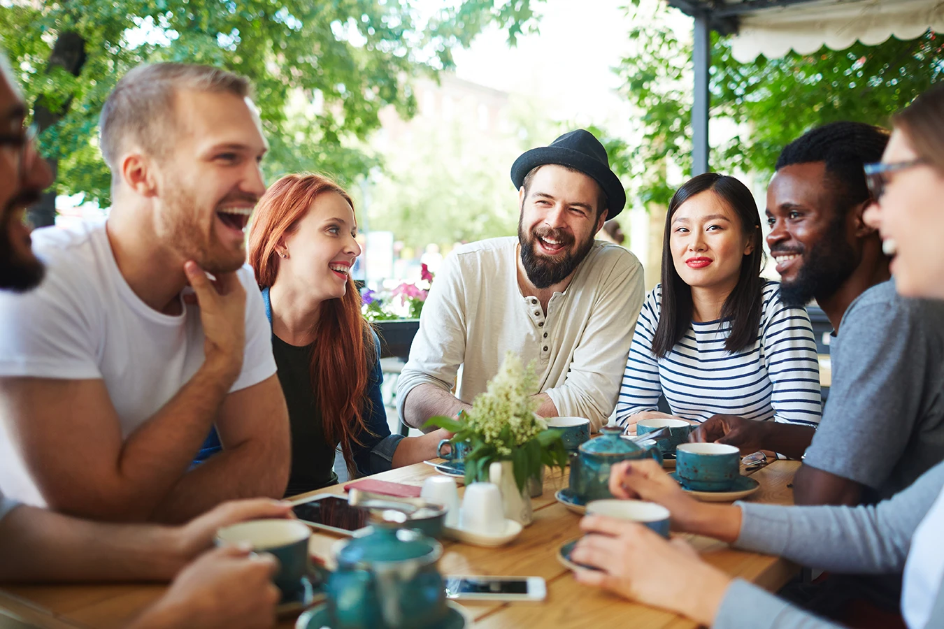 A group of seven young adults, men and women, sitting at a cafe table together talking, smiling, and laughing.