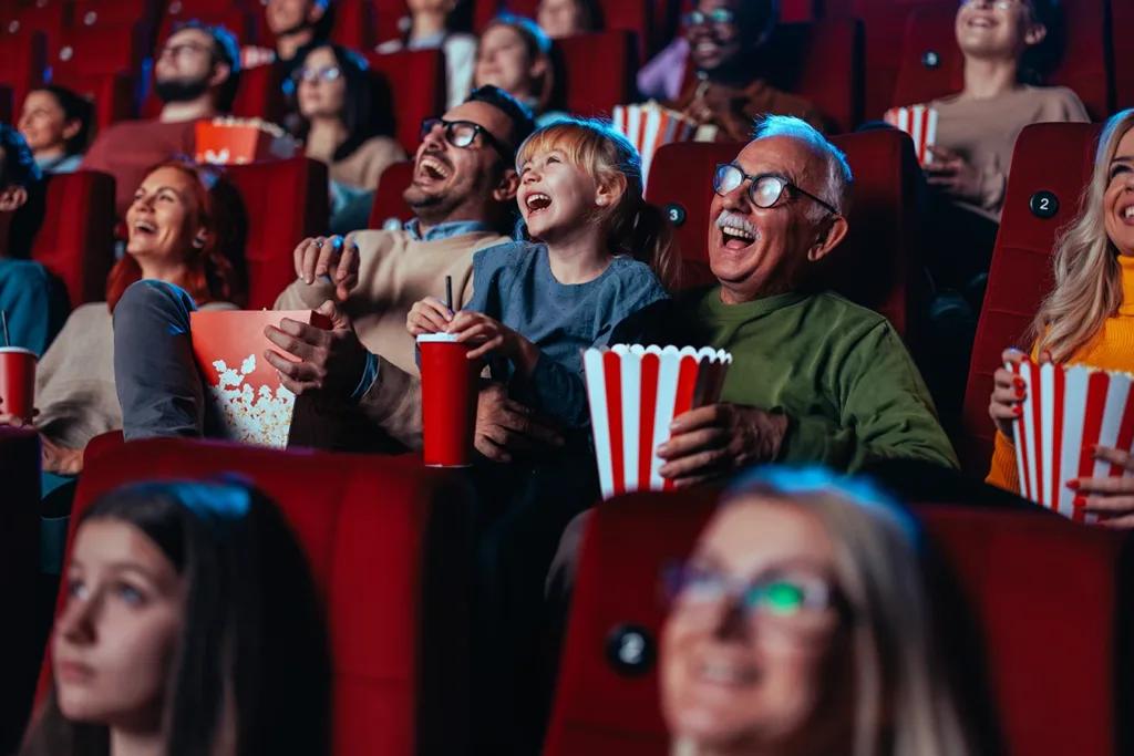 Auracast hearing aid connectivity concept: An older man (grandpa), a young child (grandchild), and a middle aged man (father), sitting in a movie theater, laughing at a funny movie on the screen