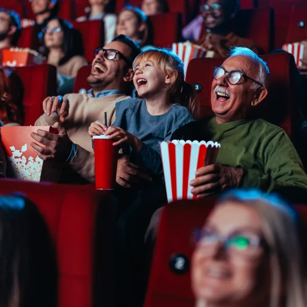 Auracast hearing aid connectivity concept: An older man (grandpa), a young child (grandchild), and a middle aged man (father), sitting in a movie theater, laughing at a funny movie on the screen