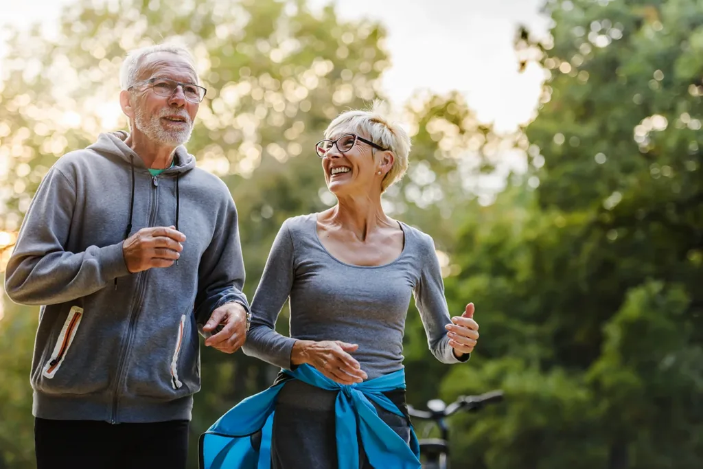 Two older adults, man and women, husband and wife, smiling while jogging side by side outside in a park. How hearing aids reduce the risk of falls concept.