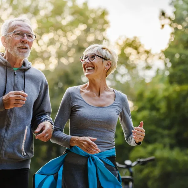 Two older adults, man and women, husband and wife, smiling while jogging side by side outside in a park. How hearing aids reduce the risk of falls concept.