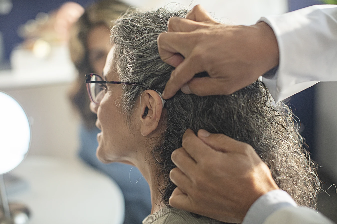 The side profile of a woman in an a hearing clinic during a hearing aid consultation, as her hearing care specialists fits a new hearing aids in her ear and powers it on