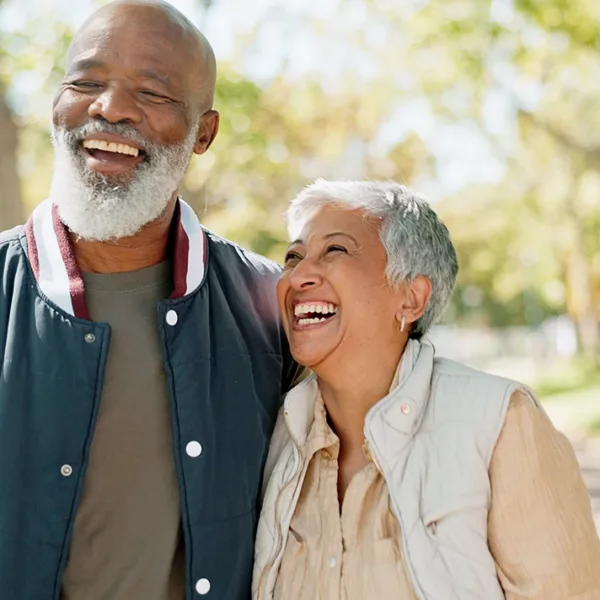A man and wife, senior couple, laughing together as the walk through a beautiful outdoor park area with lots if bright sunshine and trees in the background