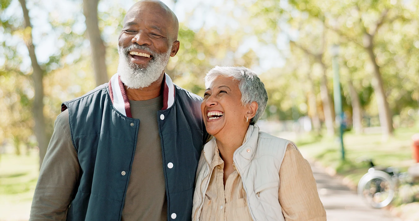 A man and wife, senior couple, laughing together as the walk through a beautiful outdoor park area with lots if bright sunshine and trees in the background