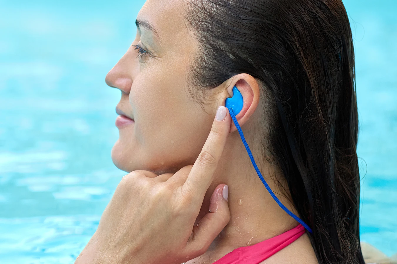 Side profile of a middle aged women with long dark hair, standing in a swimming pool, putting earplugs in to prevent swimmer's ear