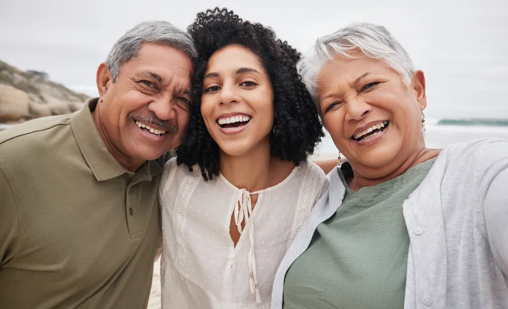 A selfie-style portrait of a young women with her older aged parents at the beach, smiling, and laughing, enjoying quality time together, symbolizing helping a loved one adjust to new hearing aids