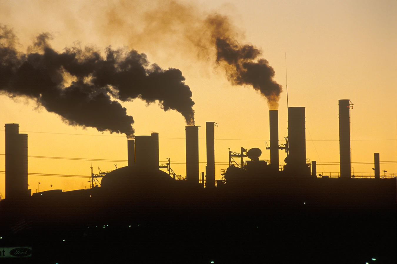 Factory smokestacks venting smoke, blocking out the sun, over a dramatic, yellow sky, symbolizing air pollution and hearing los