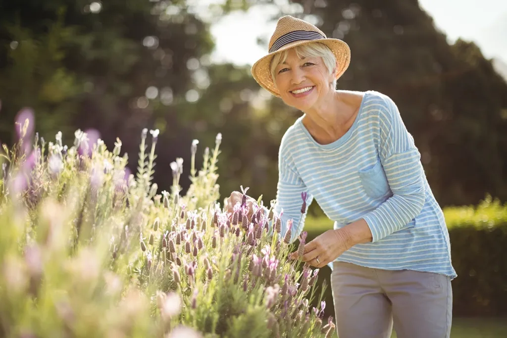 Happy older woman, smiling while tending to her lavender garden in backyard, symbolizing the many ways hearing aid use can benefit the mental health and emotional well-being of older adults.