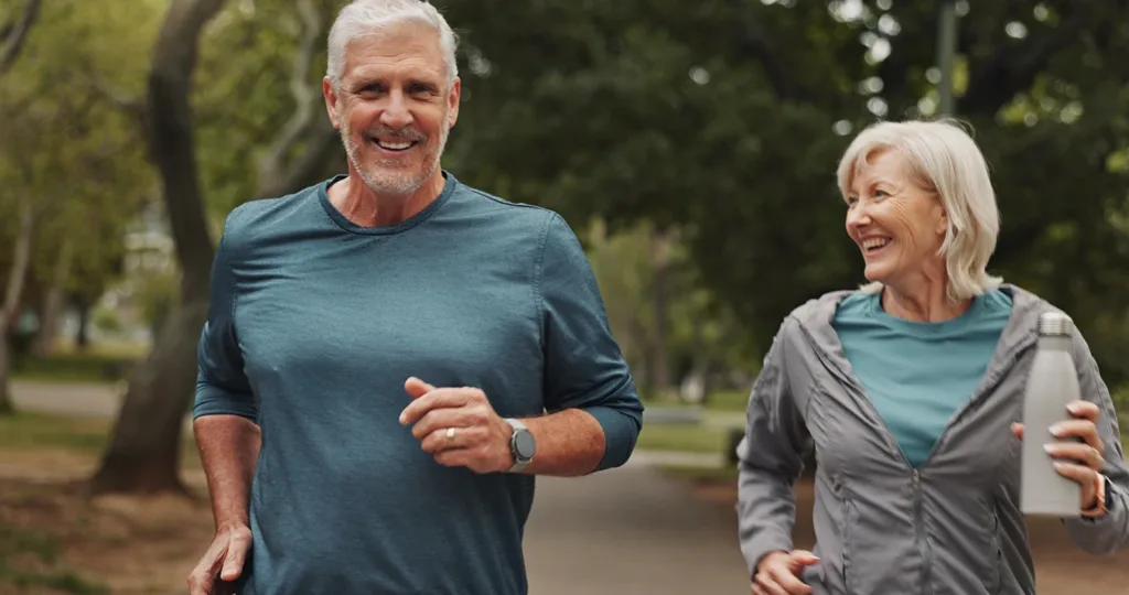 Two healthy, active, mature, senior adults, husband and wife, wearing exercise attire, jogging side, by side in a beautiful outdoor park, smiling and enjoying their workout. 