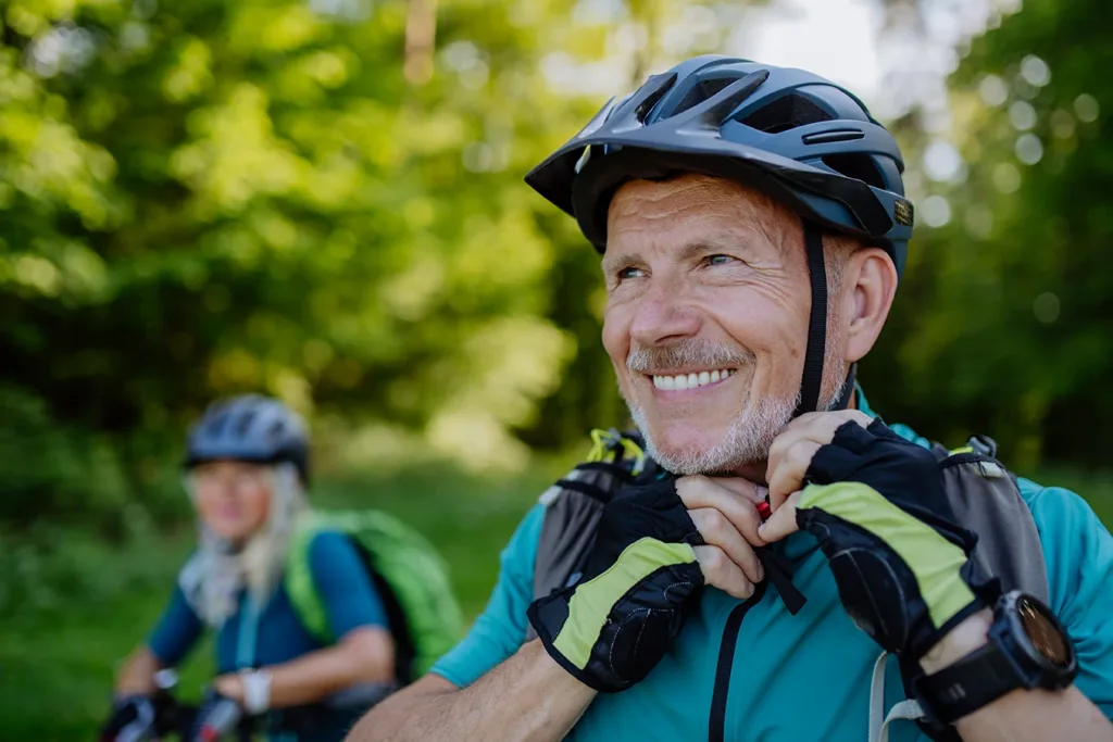 Two healthy, active, mature senior adults, husband and wife, wearing bicycle riding attire, gloves, and helmets, riding bikes outdoors in a lush green wooded area, symbolizing the importance of timely hearing care for health aging