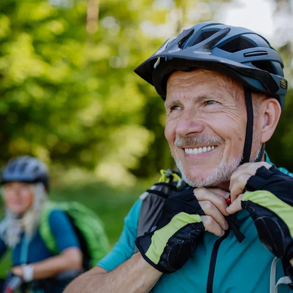 Two healthy, active, mature senior adults, husband and wife, wearing bicycle riding attire, gloves, and helmets, riding bikes outdoors in a lush green wooded area, symbolizing the importance of timely hearing care for health aging