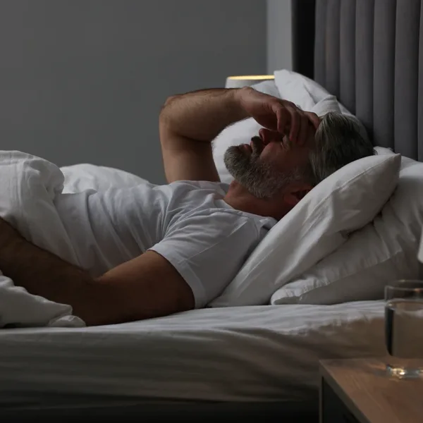Mature man laying in bed with all the lights off, feeling unwell, with his hand on his head due to a migraine.