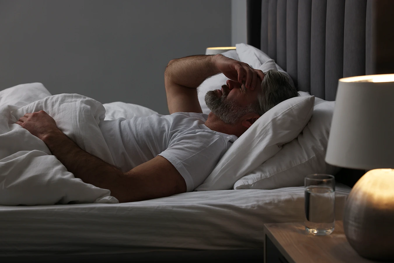 Mature man laying in bed with all the lights off, feeling unwell, with his hand on his head due to a migraine.
